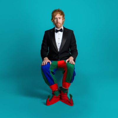 Mark Simmons is sitting on a chair against a blue background and is wearing a black tux with a bow tie and colourful patchwork pants.