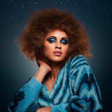 Michelle de Swarte is wearing a blue top and has bright blue eyeshadow on and she's sitting against a grey background