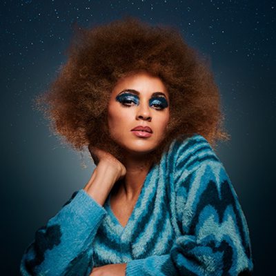 Michelle de Swarte is wearing a blue top and has bright blue eyeshadow on and she's sitting against a grey background