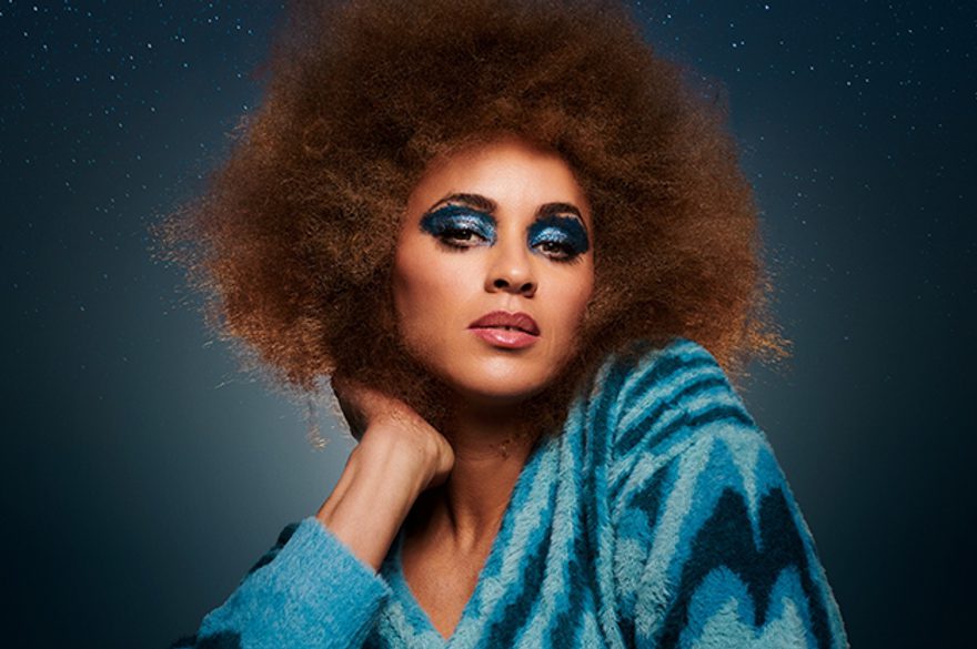 Michelle de Swarte is wearing a blue top and has bright blue eyeshadow on and she's sitting against a grey background