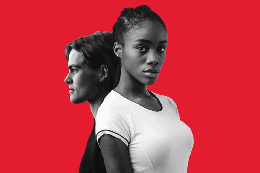 A black and white image of two young adults on a red background, a white male and a black female. The boy is positioned in profile behind the girl. She is wearing a white t-shirt and looking directly at the viewer.