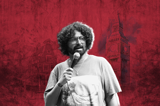 Ravi Gupta has big curly hair and is wearing glasses while holding a microphone, ready to speak or perform.