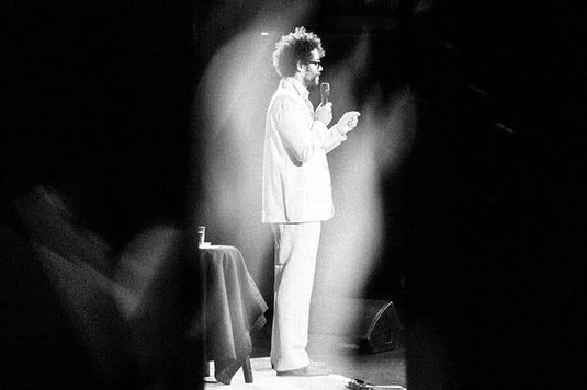 Richard Ayoade is dressed in a white suit and stands on stage, poised and ready to speak to the audience.