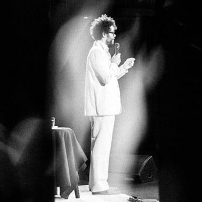 Richard Ayoade is dressed in a white suit and stands on stage, poised and ready to speak to the audience.