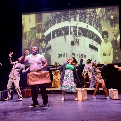 A group of people on stage dancing and signing while a picture of the Windrush ship is projected on the wall behind them