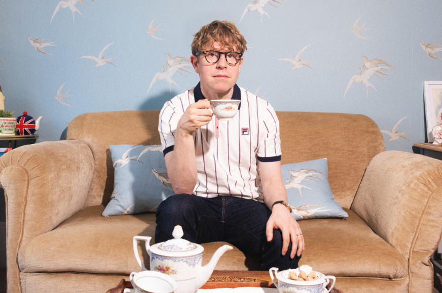 Josh Widdicombe is wearing a striped polo shirt and drinking tea. He is sat on a brown sofa with blue wallpaper with birds on behind him and a tea set on the table in front.