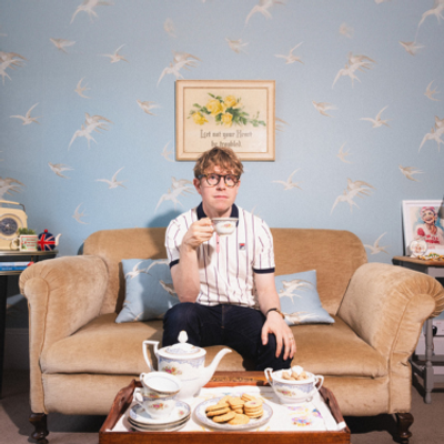 Josh Widdicombe is wearing a striped polo shirt and drinking tea. He is sat on a brown sofa with blue wallpaper with birds on behind him and a tea set on the table in front.