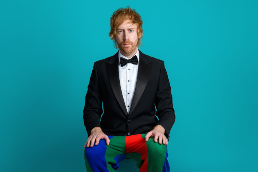 Mark Simmons is sitting on a chair against a blue background and is wearing a black tux with a bow tie and colourful patchwork pants.