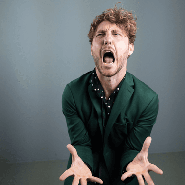 Seann Walsh is standing against a grey wall wearing a green shirt and is screaming with his hands out in front of him