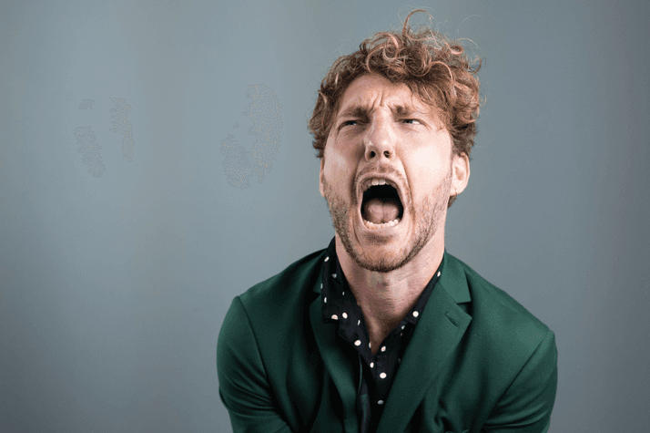 Seann Walsh is standing against a grey wall wearing a green shirt and is screaming with his hands out in front of him