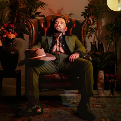 Adam Rowe is wearing a green suit and has a hat resting on his leg. He is seated in a  chair, exuding a calm and thoughtful demeanor.