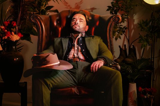 Adam Rowe is wearing a green suit and has a hat resting on his leg. He is seated in a  chair, exuding a calm and thoughtful demeanor.