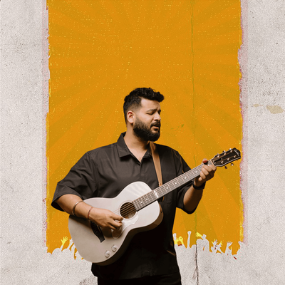 Bhuwin is a bearded musician and he is playing the guitar, expressing passion through his music. He is standing against a white wall with a splash of orange