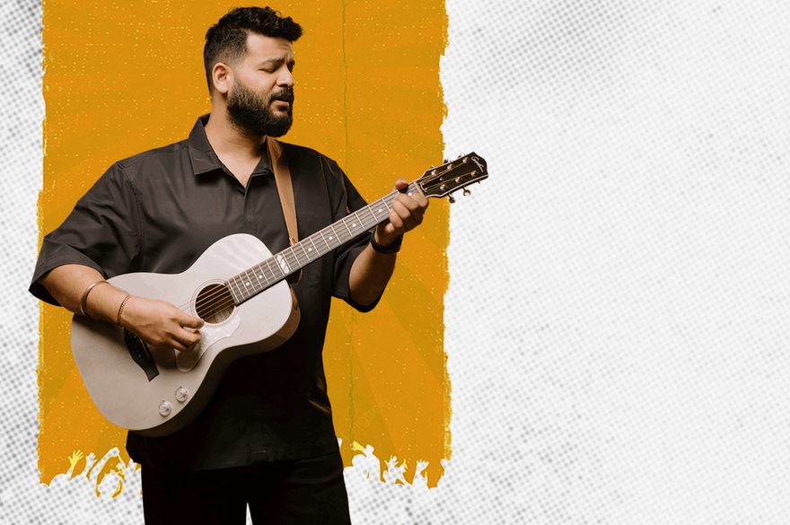 Bhuwin is a bearded musician and he is playing the guitar, expressing passion through his music. He is standing against a white wall with a splash of orange