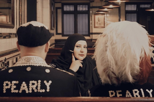 Fatiha El-Ghorri is wearing a black hijab sits and is sitting between two people wearing black hats and jackets with “PEARLY” spelled out in white beads, inside a diner-style restaurant.
