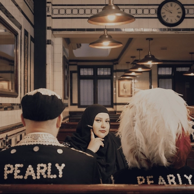 Fatiha El-Ghorri is wearing a black hijab sits and is sitting between two people wearing black hats and jackets with “PEARLY” spelled out in white beads, inside a diner-style restaurant.