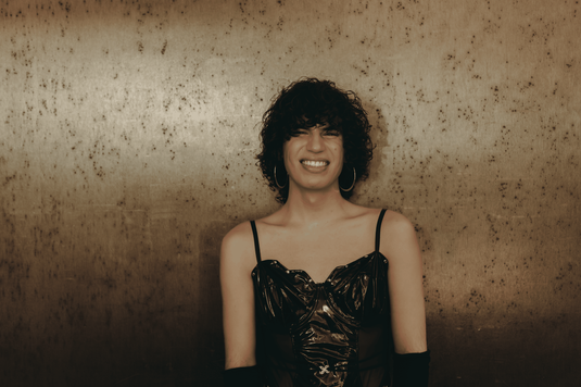 Jacob Alon has curly dark hair wearing a black glossy spaghetti-strap top and long black gloves, standing against a textured brown wall.
