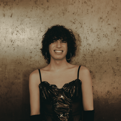 Jacob Alon has curly dark hair wearing a black glossy spaghetti-strap top and long black gloves, standing against a textured brown wall.