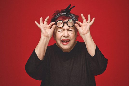 Jo Brand has red hair and a black headband holds a pair of round glasses in front of her face while squinting against a bright red background.