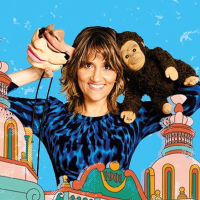 Nina Conti has short brown hair and is holding a hand puppet in one hand and has a plush monkey draped over the opposite shoulder. They wear a blue-and-black patterned top and stand against a bright blue illustrated background featuring colorful, whimsical architectural elements.