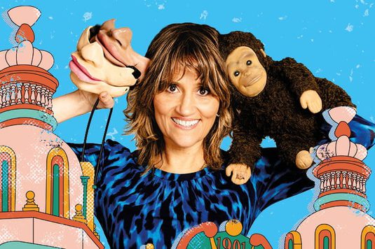 Nina Conti has short brown hair and is holding a hand puppet in one hand and has a plush monkey draped over the opposite shoulder. They wear a blue-and-black patterned top and stand against a bright blue illustrated background featuring colorful, whimsical architectural elements.