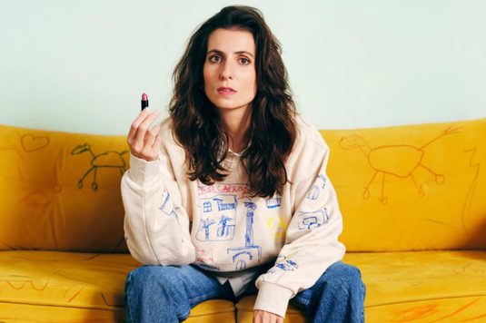 A woman sits on a bright yellow sofa covered in childlike drawings, holding a lipstick and looking at the camera. Toys and coloured pencils are scattered on a play mat at her feet, with a single shoe placed to one side.