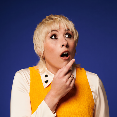 Masie Adam is wearing a white shirt and a yellow jumper on top. She has a surprised look, her eyebrows raised and mouth agape, indicating a moment of unexpected revelation.