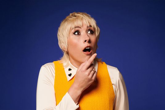 Masie Adam is wearing a white shirt and a yellow jumper on top. She has a surprised look, her eyebrows raised and mouth agape, indicating a moment of unexpected revelation.