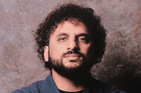 Nish Kumar has black curly hair and a beard. He is wearing a dark blue shirt and is standing confidently against a brown wall, posing for a photograph.