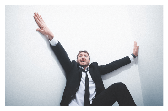 Seann Walsh in a suit is sat against a white background with arms outstretched and a pained expression