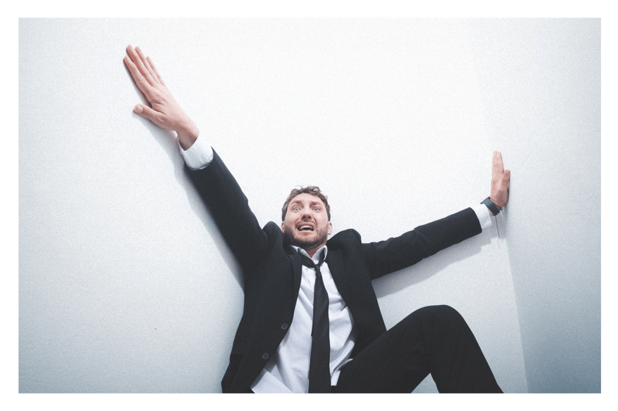 Seann Walsh in a suit is sat against a white background with arms outstretched and a pained expression