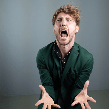 Seann Walsh is standing against a grey wall wearing a green shirt and is screaming with his hands out in front of him