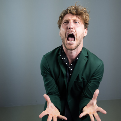 Seann Walsh is standing against a grey wall wearing a green shirt and is screaming with his hands out in front of him