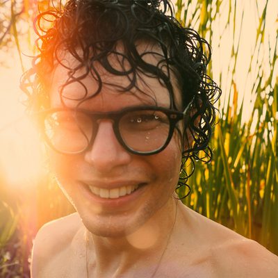 Simon Amstell is wearing glasses and ha no shirt on and is seen in a field, with tall grass and a sunny sky creating a serene outdoor scene.