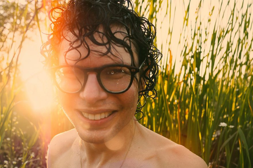 Simon Amstell is wearing glasses and ha no shirt on and is seen in a field, with tall grass and a sunny sky creating a serene outdoor scene.