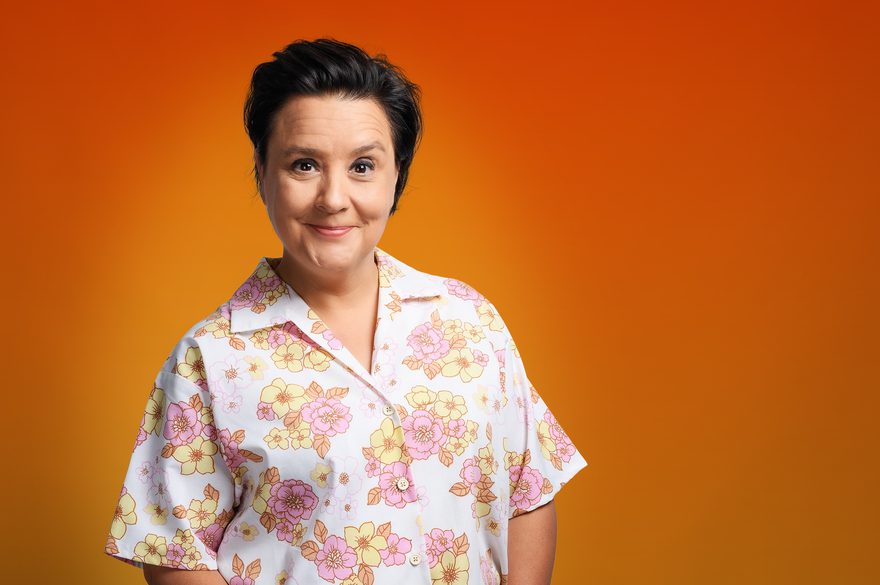 Susan Calman has short dark hair and is wearing a floral shirt in soft pink and yellow tones. Susan Calman is smiling straight at the camera, The background is a vibrant orange, creating a warm and cheerful atmosphere.
