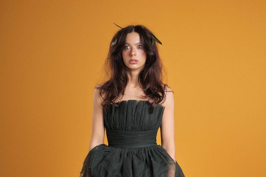 Abi Clarke has long dark brown hair and is standing against a warm orange background, wearing a strapless black dress with a pleated bodice and full, gathered skirt. Green leaves are tucked into her hair, and she looks directly at the camera with a neutral expression.