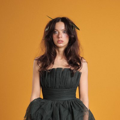 Abi Clarke has long dark brown hair and is standing against a warm orange background, wearing a strapless black dress with a pleated bodice and full, gathered skirt. Green leaves are tucked into her hair, and she looks directly at the camera with a neutral expression.