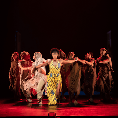 A group of eight dancers perform on a dark theater stage under dramatic red lighting. The central dancer wears a bright yellow patterned outfit, while the surrounding dancers wear flowing brown, rust, and cream-colored costumes. They extend their arms outward in a coordinated formation, with the glossy stage floor reflecting the red light.