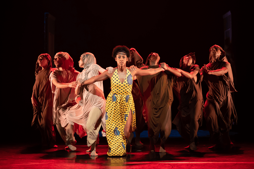 A group of eight dancers perform on a dark theater stage under dramatic red lighting. The central dancer wears a bright yellow patterned outfit, while the surrounding dancers wear flowing brown, rust, and cream-colored costumes. They extend their arms outward in a coordinated formation, with the glossy stage floor reflecting the red light.