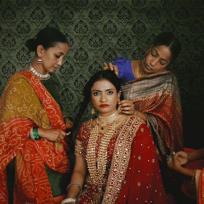 A bride in a red and gold sari sits while three women in colorful traditional Indian clothing help arrange her hair, jewelry, and sari. The group is posed against an ornate dark green patterned backdrop.