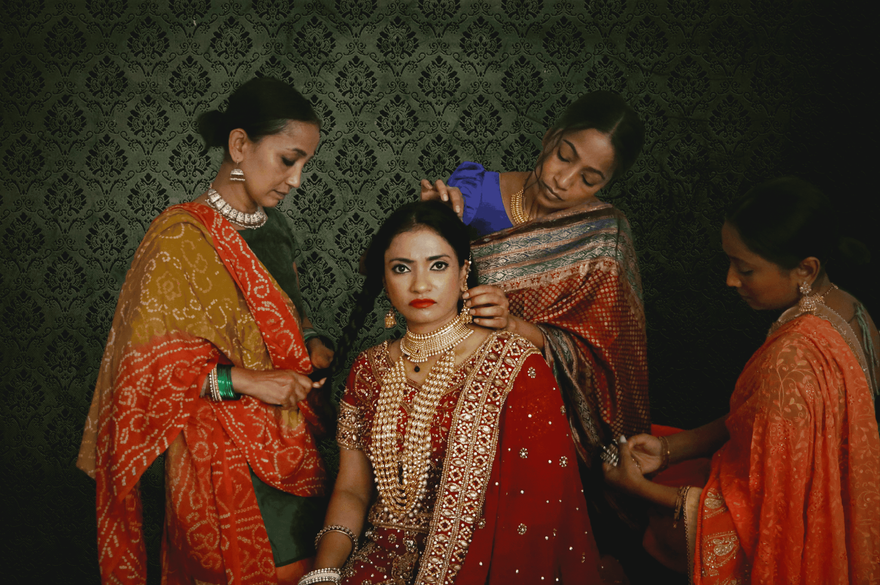 A bride in a red and gold sari sits while three women in colorful traditional Indian clothing help arrange her hair, jewellery, and sari. The group is posed against an ornate dark green patterned backdrop.