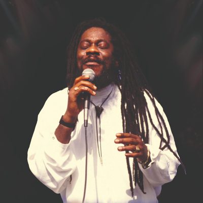 Dennis Brown has long dreadlocks and is wearing a white shirt as he performs on stage with eyes closed, holding a microphone.