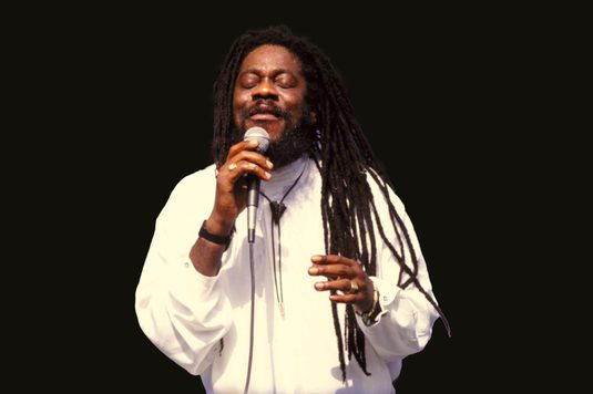 Dennis Brown has long dreadlocks and is wearing a white shirt as he performs on stage with eyes closed, holding a microphone.