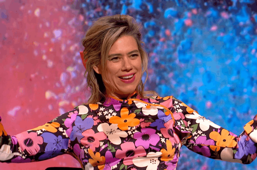 A colourful close-up portrait of a Lou Sanders smiling with short, swept-back brown hair, wearing a vibrant floral-patterned top. She is looking towards the camera against a bright, blurred pink and blue background.