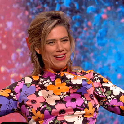 A colourful close-up portrait of a Lou Sanders smiling with short, swept-back brown hair, wearing a vibrant floral-patterned top. She is looking towards the camera against a bright, blurred pink and blue background.