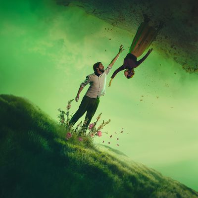 Two figures stand suspended against a green misty landscape. A man reaches upwards towards a woman who is floating upside down above him. Their outstretched hands nearly meet. Behind them, a hazy green sky or mist creates an ethereal atmosphere, with dark, textured rock formations framing the upper edge of the image