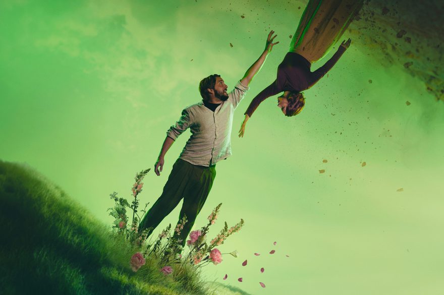 Two figures stand suspended against a green misty landscape. A man reaches upwards towards a woman who is floating upside down above him. Their outstretched hands nearly meet. Behind them, a hazy green sky or mist creates an ethereal atmosphere, with dark, textured rock formations framing the upper edge of the image