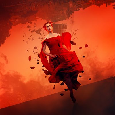 Looking fearful and frantic, a woman in a red dress runs towards the frame. The landscape behind her is red and smokey and the grand building behind her is upside down. The building falls and crumbles and she is surrounded by debris from the destruction as she runs.