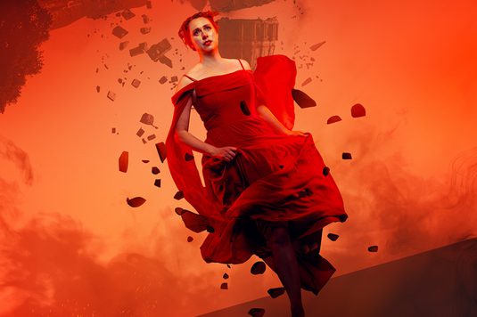 Looking fearful and frantic, a woman in a red dress runs towards the frame. The landscape behind her is red and smokey and the grand building behind her is upside down. The building falls and crumbles and she is surrounded by debris from the destruction as she runs.